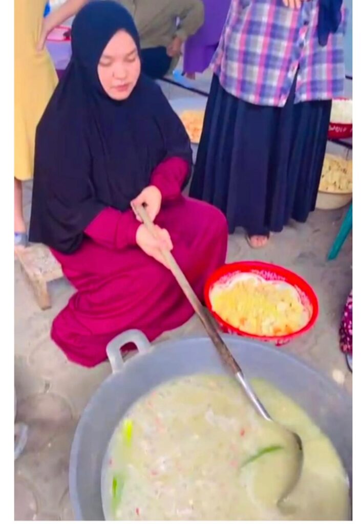 Whatsapp image 20260616 at 16 06 19 catrawarta Woman wearing a black hijab and magenta dress stirs a large pot of soupporridge with a long wooden paddle at an outdoor market